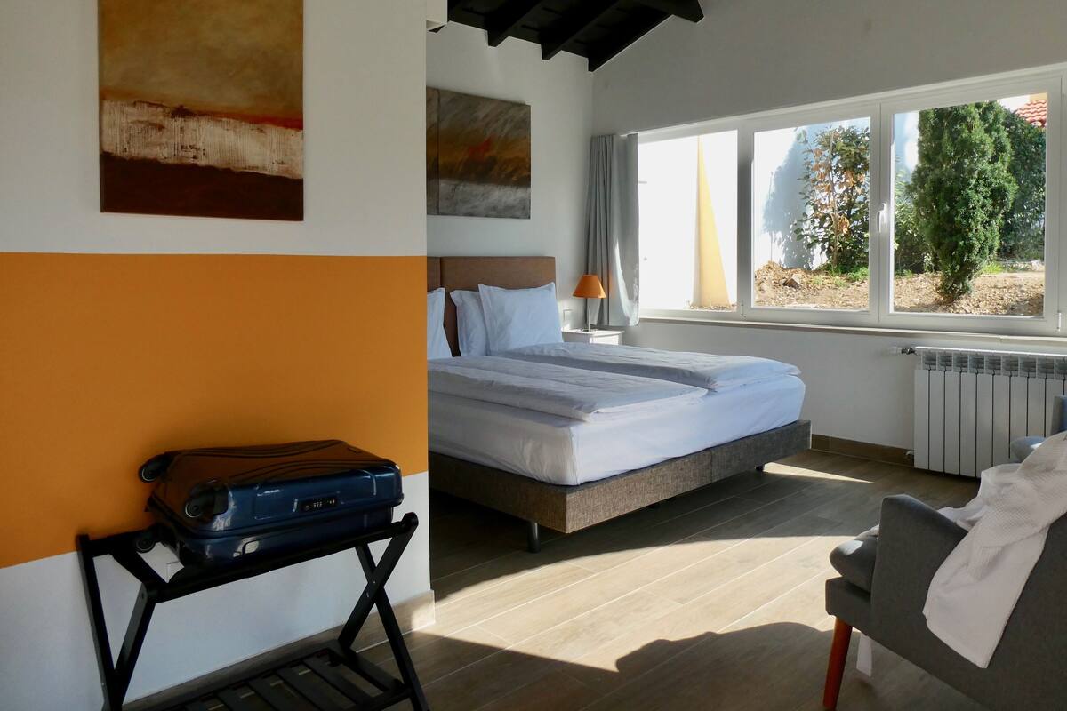 A bedroom with a bed, a nightstand, a lamp, a suitcase, and a view of trees through the window.