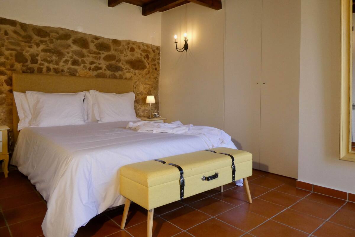 A bedroom with a stone wall, a bed with white linens, a bedside lamp, and a suitcase bench.