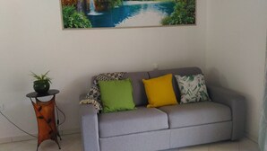 Un canapé gris agrémenté de coussins verts et jaunes, une petite table avec une plante et un tableau représentant une cascade au mur.