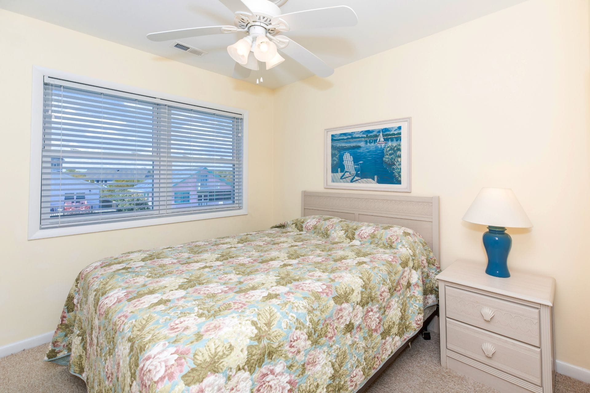 A bedroom with a bed, a nightstand with a lamp, a ceiling fan, a window with blinds, and a painting on the wall.