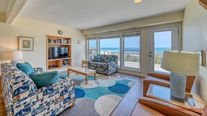 A living room with a sofa, armchair, coffee table, TV, bookshelf, and a view of the ocean through large windows. - Periwinkle - Oceanfront 3 BR, 2.5 BA home in Surf City!  Dog Friendly! (Surf City)