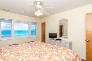 A bedroom with a bed, a television, a ceiling fan, a window with a view of the ocean, and a door.
