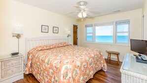Ein Schlafzimmer mit Bett, Deckenventilator, Fenster mit Meerblick, Nachttisch mit Lampe und Fernseher auf einem Schrank.