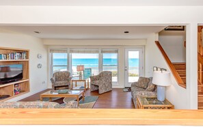 A living room with a television, bookshelves, patterned furniture, and a view of the ocean through large windows.