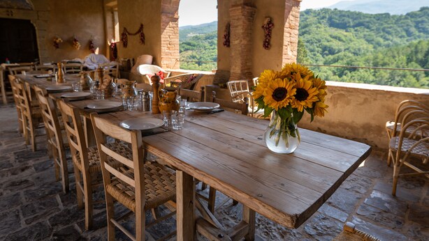 A rustic dining area with wooden tables and chairs, a vase of sunflowers, and a view of a forested landscape through an arched window. - Medieval Tuscan Castle  with 8 bedrooms for 18 guests (Seggiano)