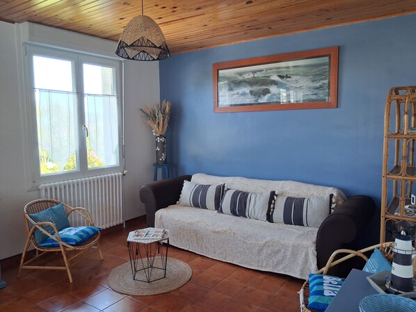 A room with a blue wall, a white sofa with striped pillows, a wicker chair, a small table, and a framed picture of a seascape.
