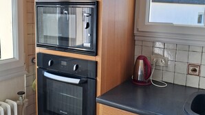 A kitchen with wooden cabinets, a built-in oven, a microwave, and a kettle on the countertop. - HOUSE NEAR ROSAIRES BEACH (Plérin)