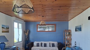 A room with a wooden ceiling, a sofa with striped cushions, a small table, a wicker lamp, and a bookshelf. - HOUSE NEAR ROSAIRES BEACH (Plérin)