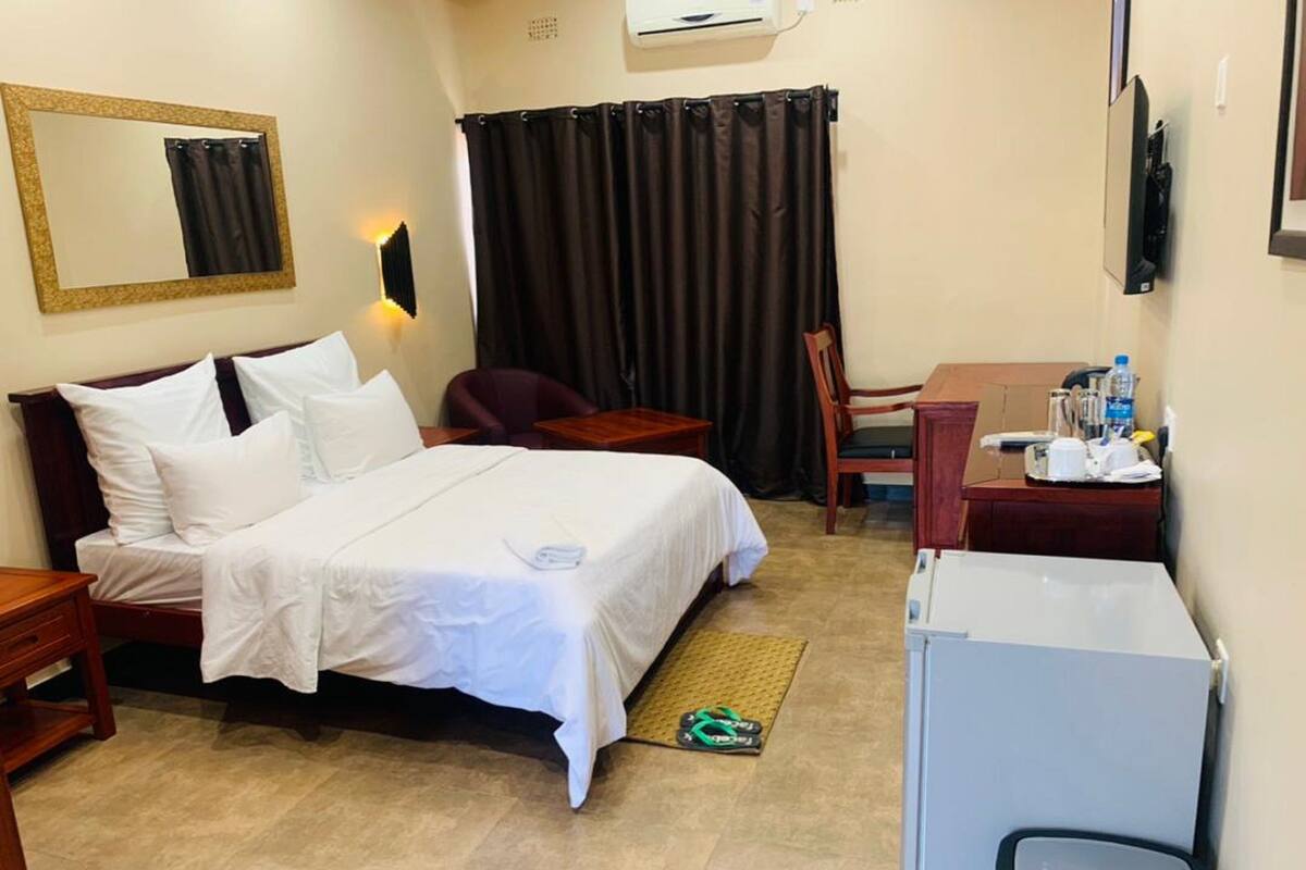 A hotel room with a bed, a desk, a chair, a mirror, a window with curtains, and a refrigerator.