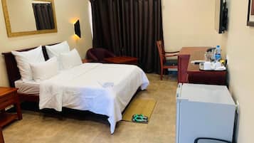 A hotel room with a bed, a desk, a chair, a mirror, a window with curtains, and a refrigerator.