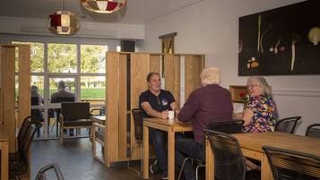 Een eethoek binnen met houten tafels en stoelen, grote ramen en een schilderij aan de muur.