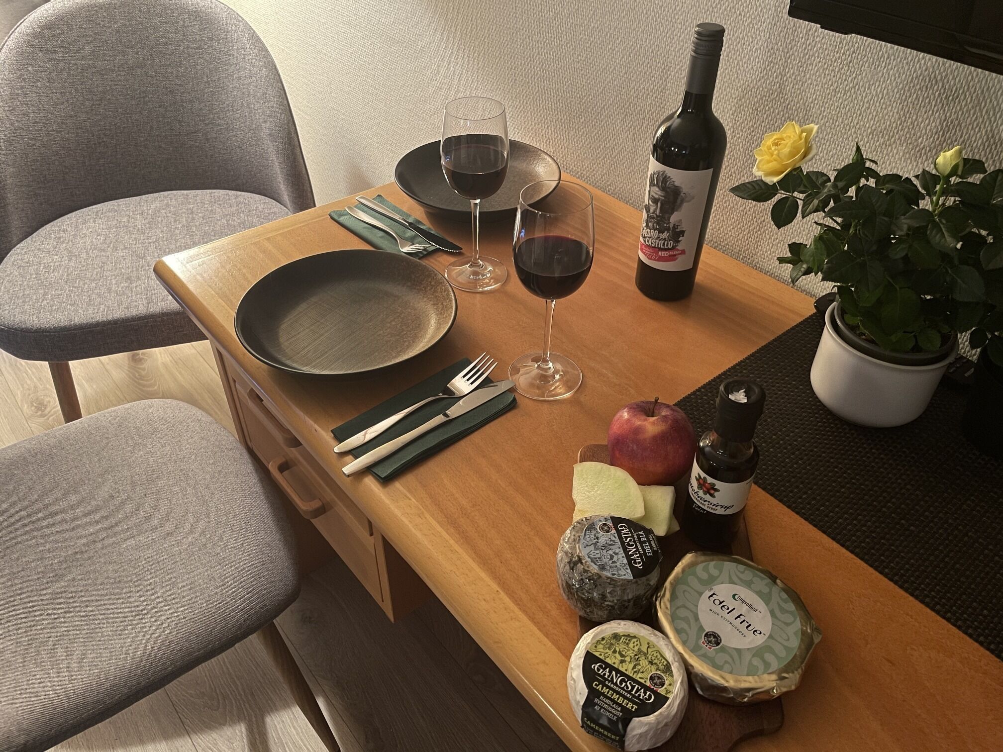A dining table with a plate, fork, knife, wine glasses, a bottle of wine, an apple, cheese, and a potted plant.