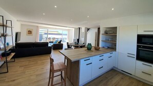 Una cocina moderna con armarios blancos, encimera de madera y horno empotrado. Hay una zona de comedor con una mesa y sillas, y un sofá negro en la sala de estar.