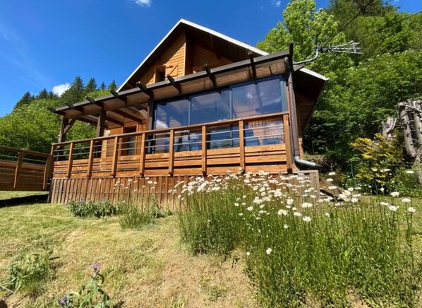 Una cabaña de madera con amplios ventanales, rodeada por un paisaje natural con flores silvestres y árboles.