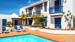 A poolside area with lounge chairs, a swimming pool, and a building with blue balconies and white walls. - Apartment 4 Small block of 6 apartments, a stones through from Marina Rubicon (Yaiza)