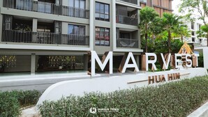 A multi-story hotel with balconies, a sign reading "MARYVEST HUA HIN," and a garden area with palm trees.