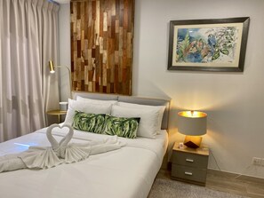 A neatly made bed with white linens and a towel arranged in a swan shape, a wooden headboard, a nightstand with a lamp, and a framed painting on the wall.