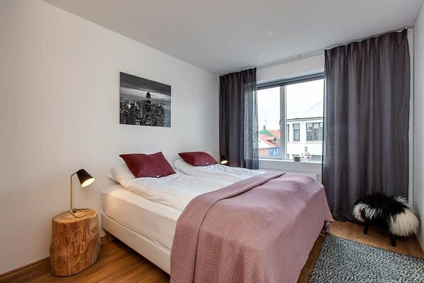 Un dormitorio moderno con una cama, un cuadro de un paisaje urbano, una mesita de noche y una ventana con cortinas.