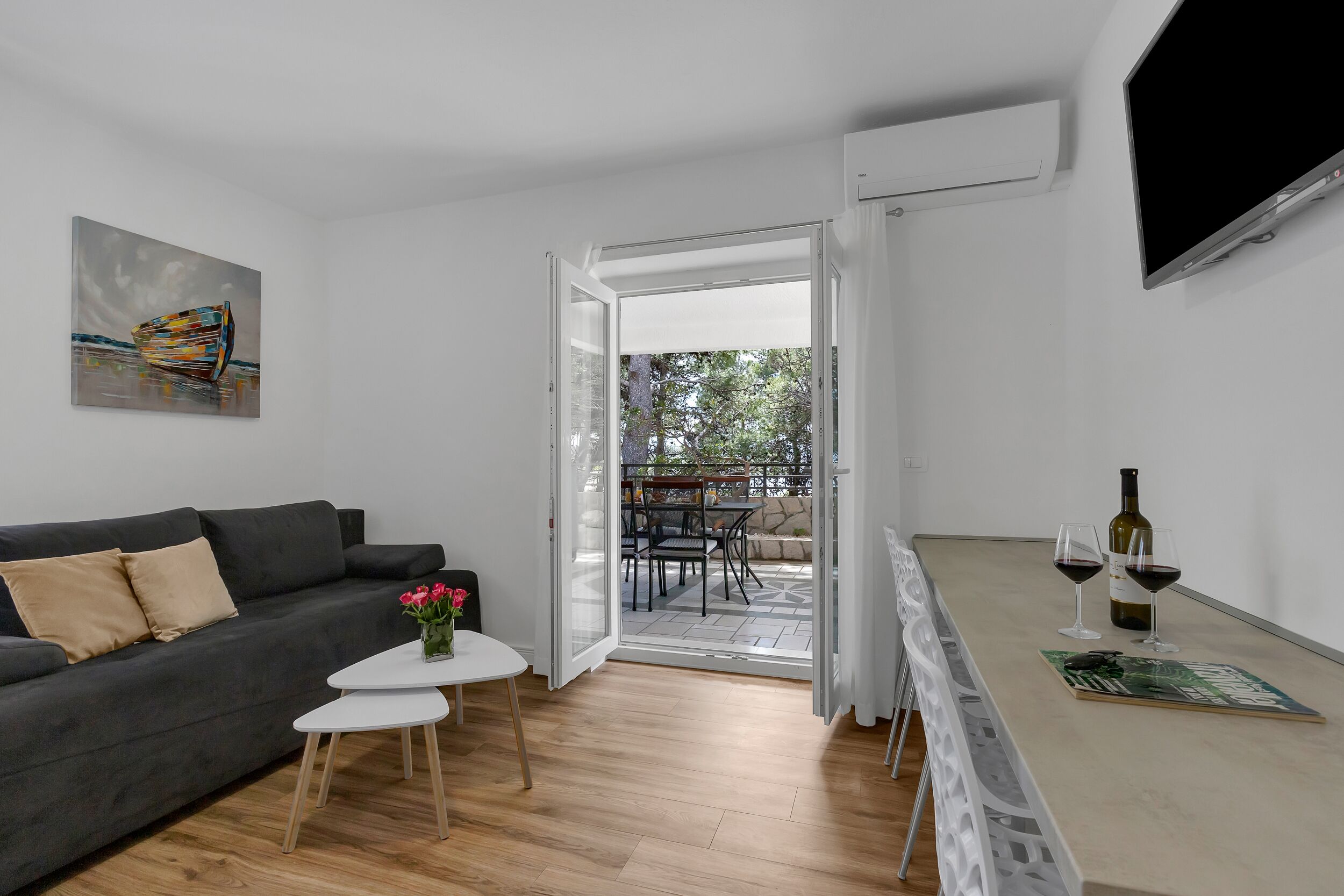 A modern living room with a grey sofa, a small white table with a vase of flowers, and a dining area with a table and chairs.