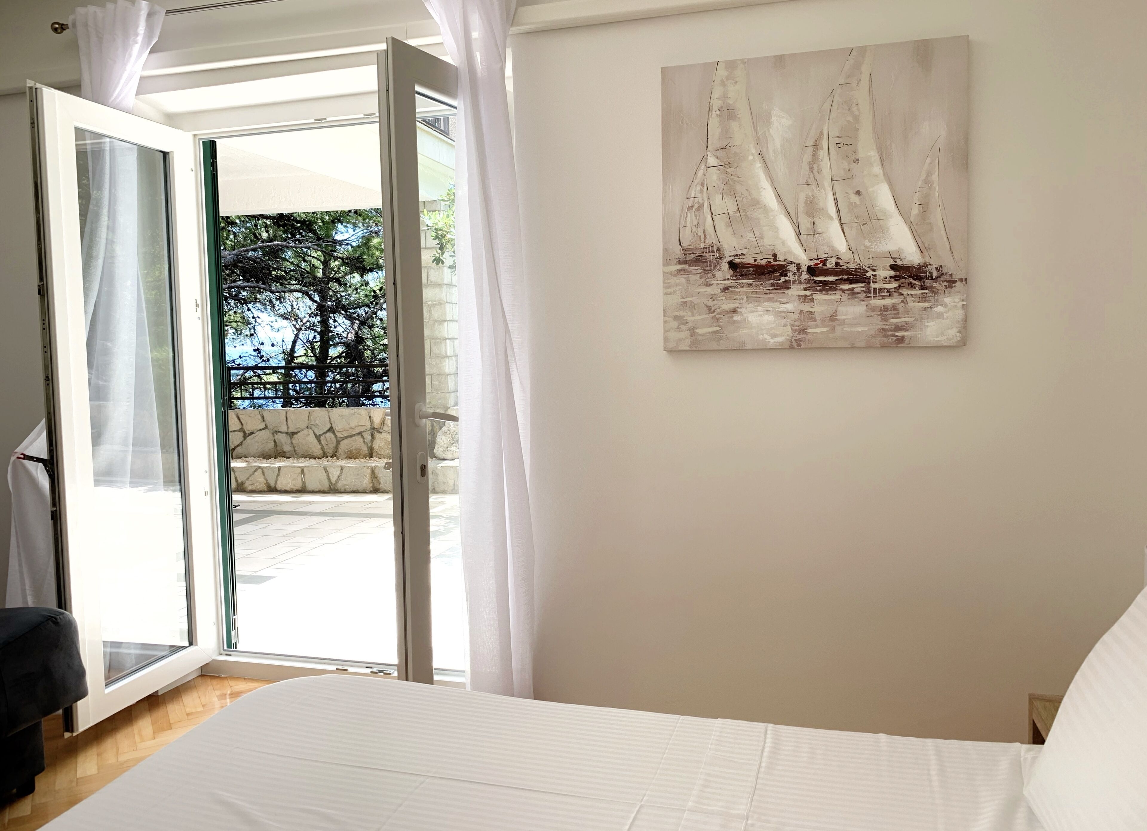 Un dormitorio con cama, un cuadro de veleros y vistas a un espacio exterior con pared de piedra y vegetación a través de una puerta abierta.