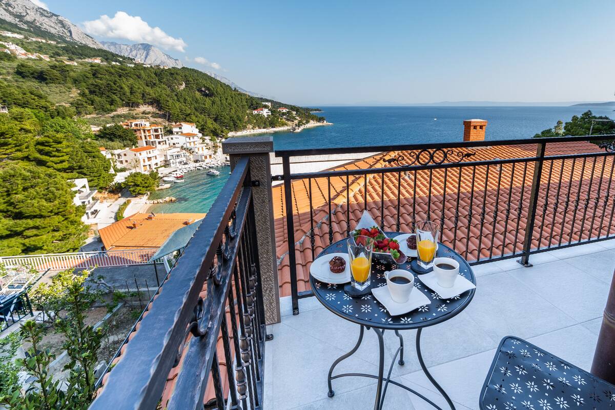 Un balcon avec une table préparée pour le petit-déjeuner, offrant une vue sur une ville côtière et la mer.