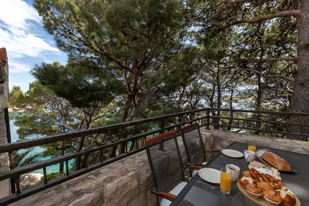 Un espace de restauration en plein air avec une table dressée pour le petit-déjeuner, offrant une vue panoramique sur des arbres et une étendue d’eau.