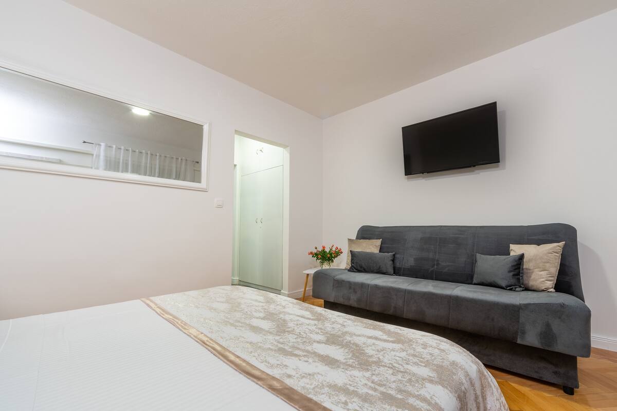 Une chambre à coucher avec un lit, un canapé gris, un téléviseur fixé au mur et une petite table d’appoint sur laquelle se trouve un vase.