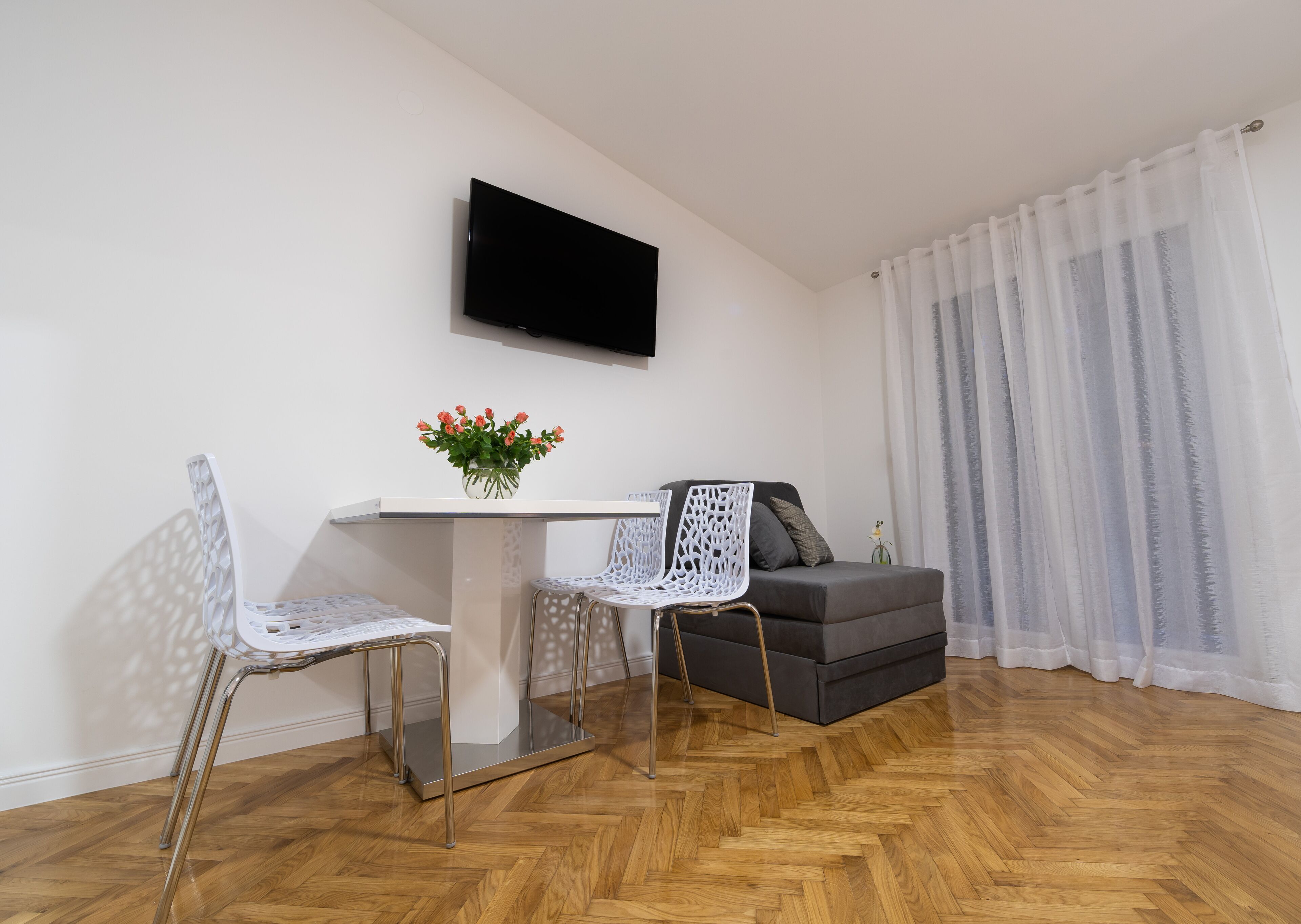 A modern living room with a wooden herringbone floor, a white table with a vase of flowers, two white chairs, a grey sofa, and a flat-screen TV mounted on the wall.