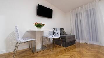 A modern living room with a wooden herringbone floor, a white table with a vase of flowers, two white chairs, a grey sofa, and a flat-screen TV mounted on the wall.