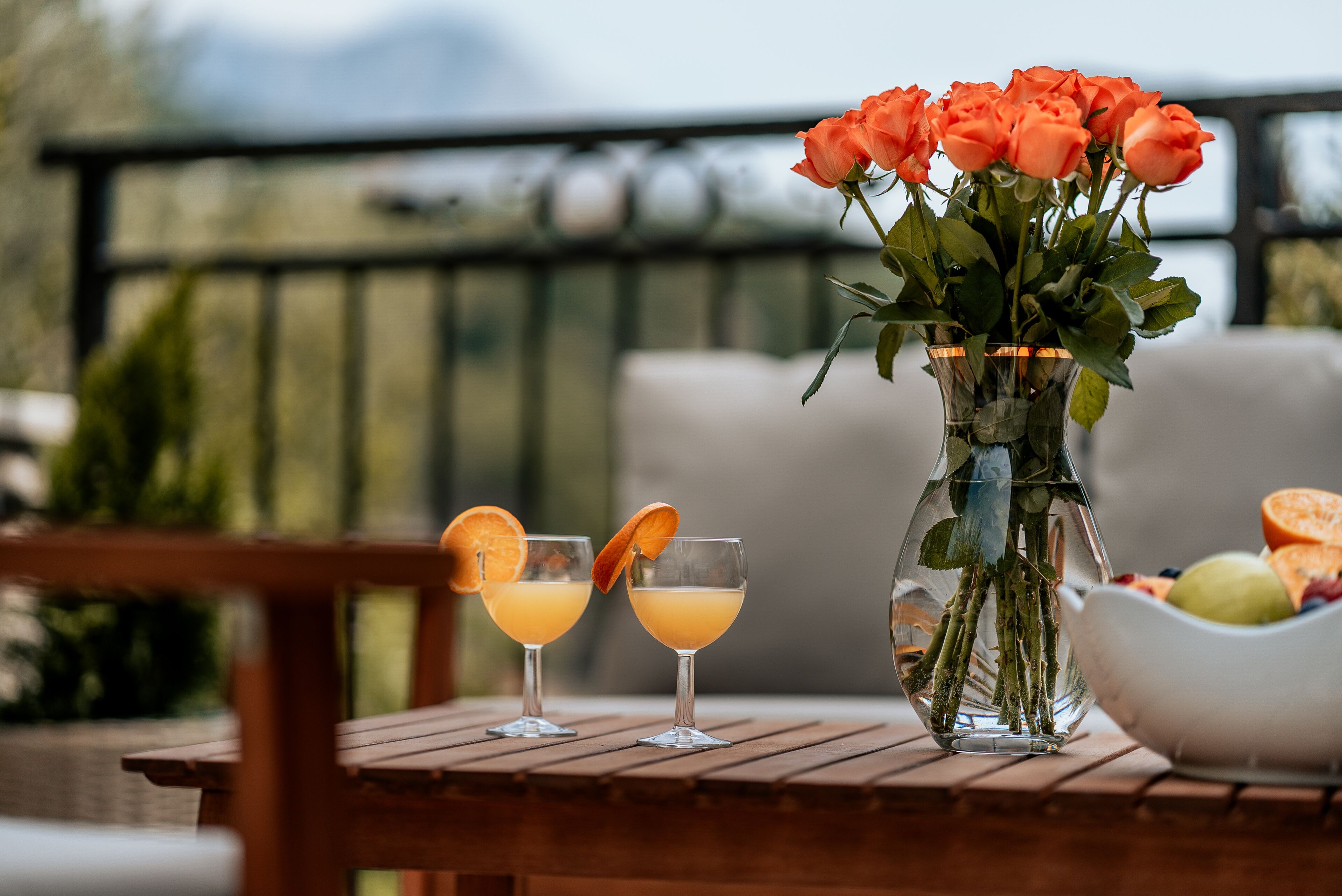 Una mesa de madera con dos vasos de jugo de naranja, un jarrón con rosas naranjas y un cuenco de fruta.