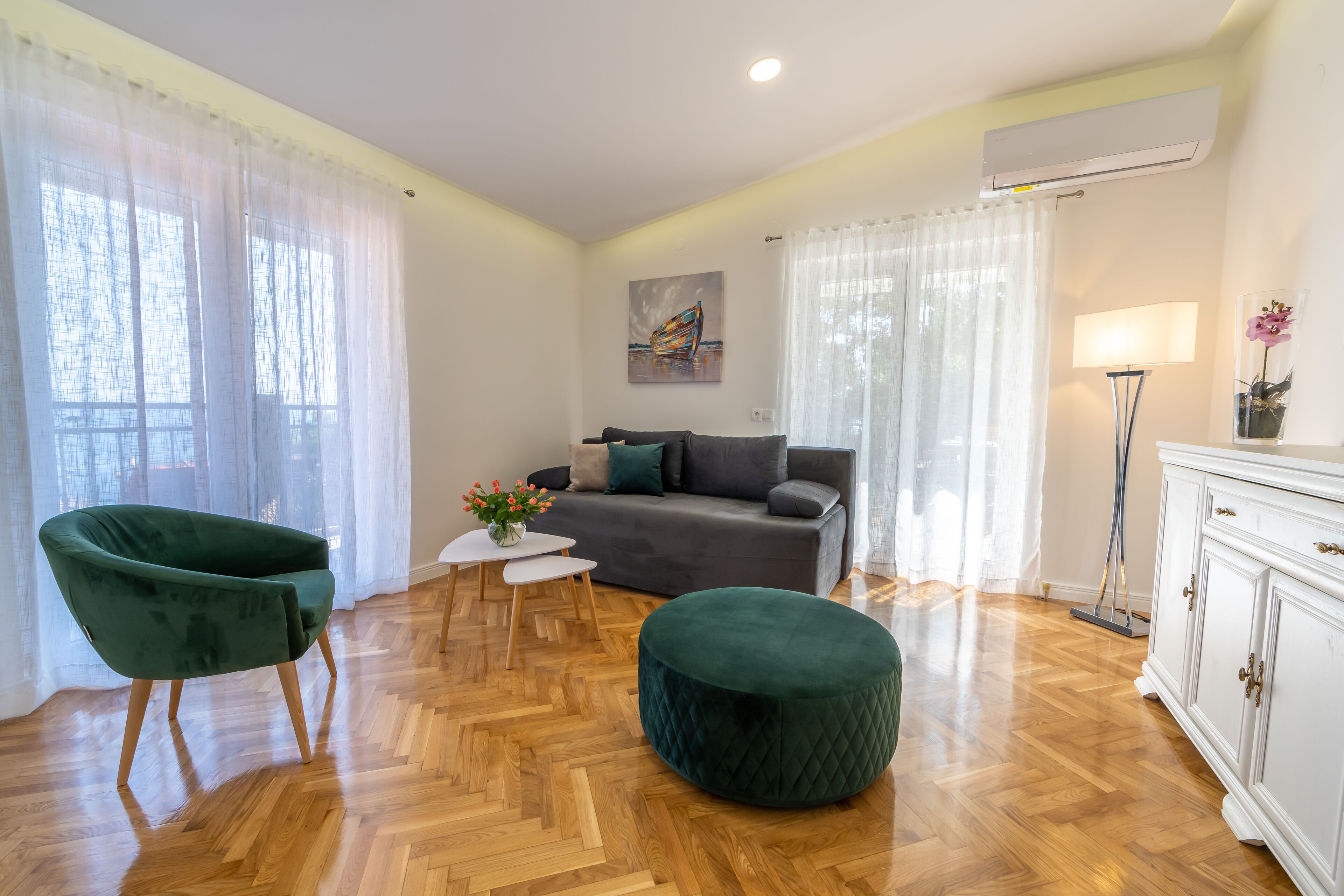 A living room with a sofa, a green armchair, a round ottoman, a small table with flowers, and a lamp.