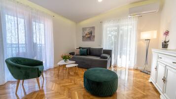 A living room with a sofa, a green armchair, a round ottoman, a small table with flowers, and a lamp.