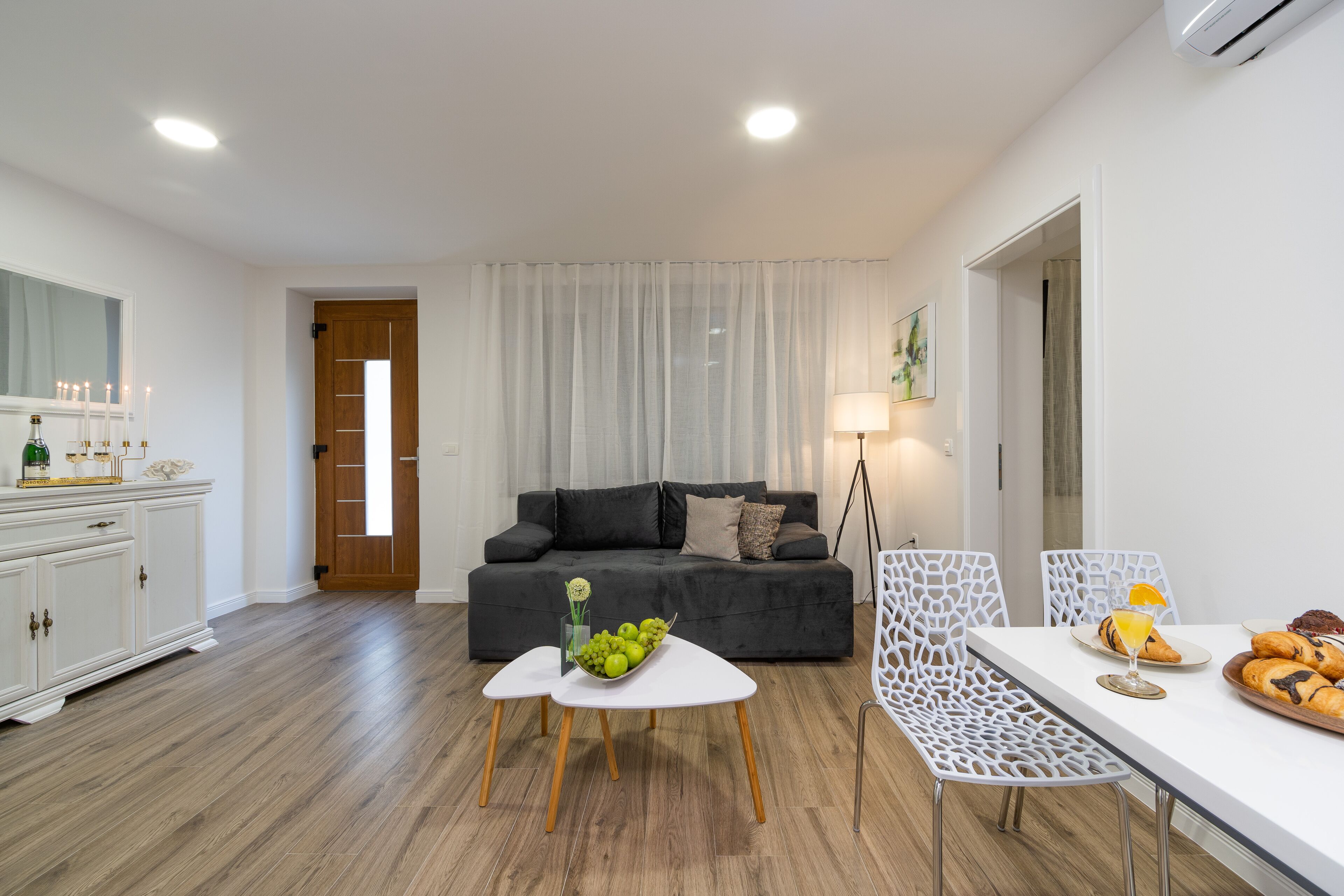 A modern living room with a dark grey sofa, white dining table, and chairs, a white cabinet, and a wooden floor.