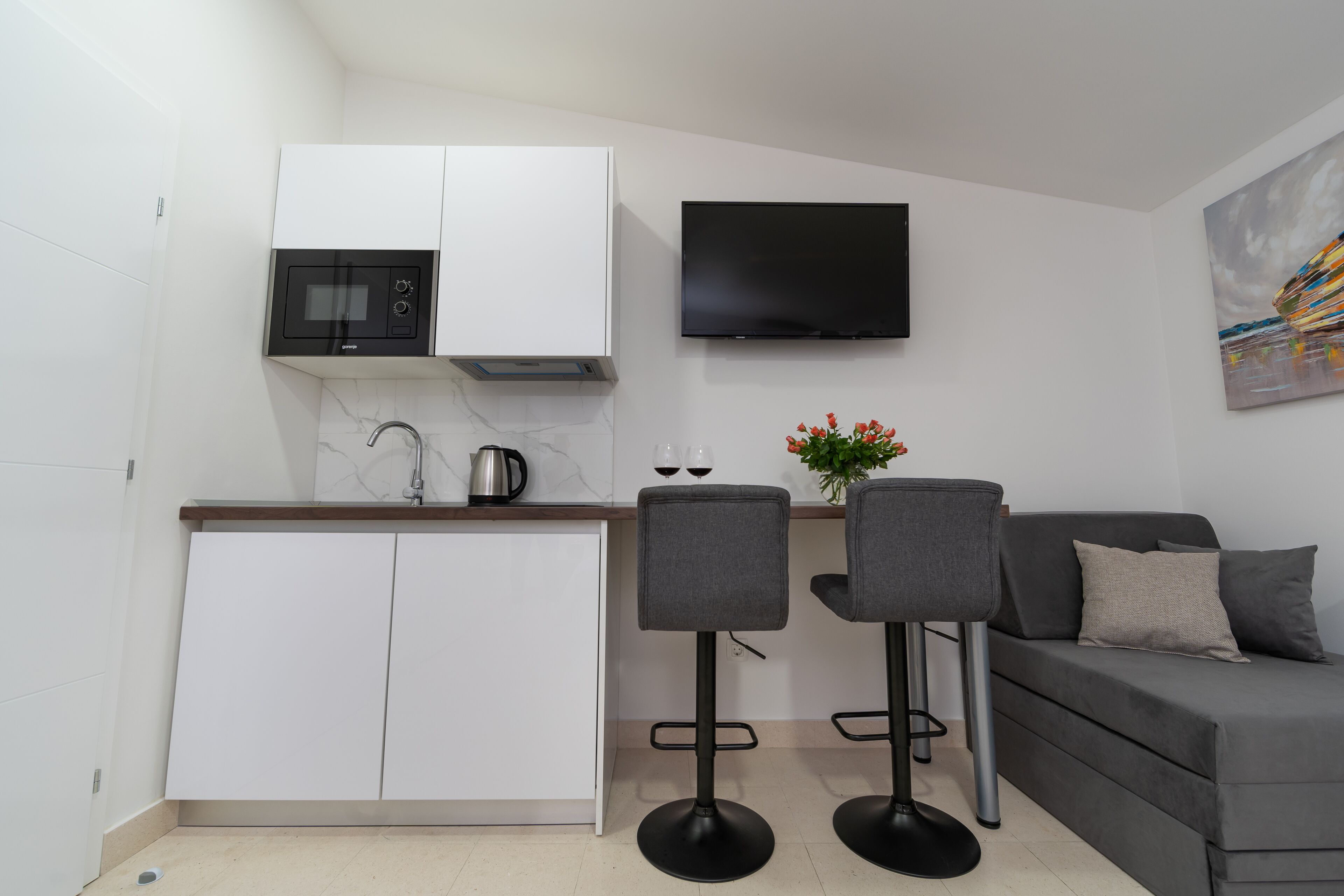 Un espacio habitable compacto con kitchenette, un televisor de pantalla plana, un sofá y dos taburetes tipo bar.
