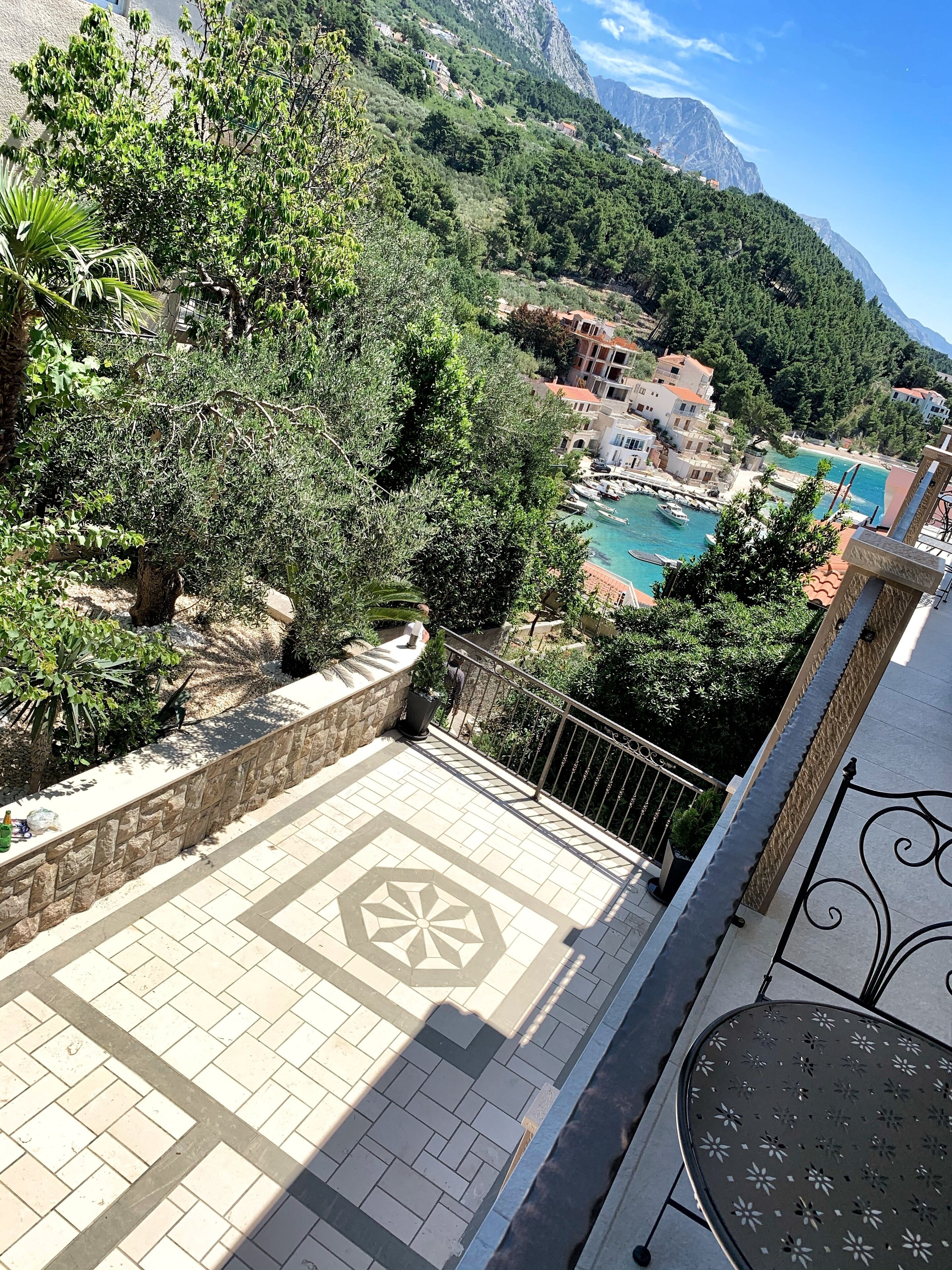 Um terraço com piso de azulejos, parede de pedra e vista para uma área montanhosa com edifícios e um corpo d'água.
