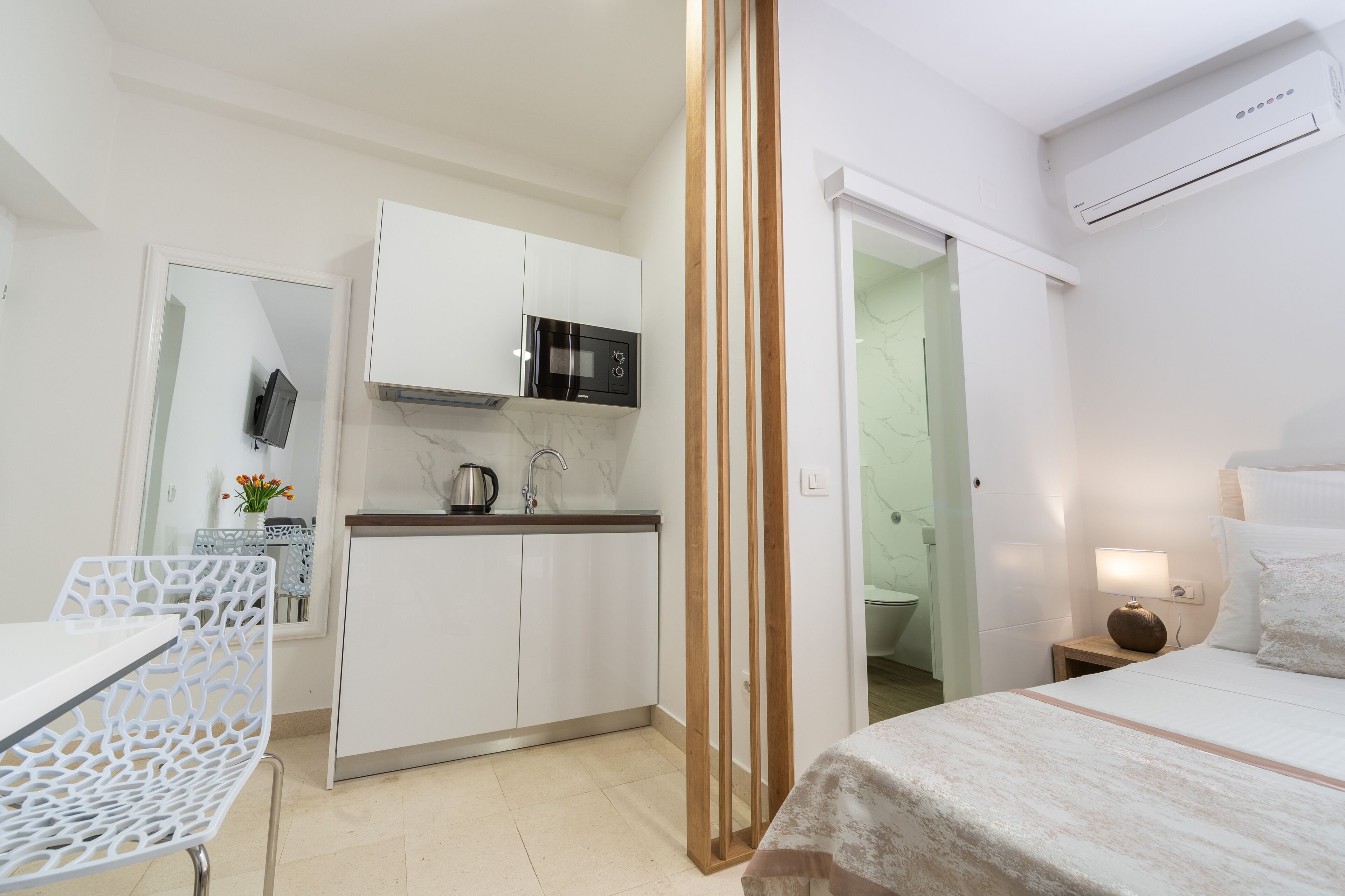 A compact hotel room with a kitchenette, a bed, a bedside table, and a mirror.