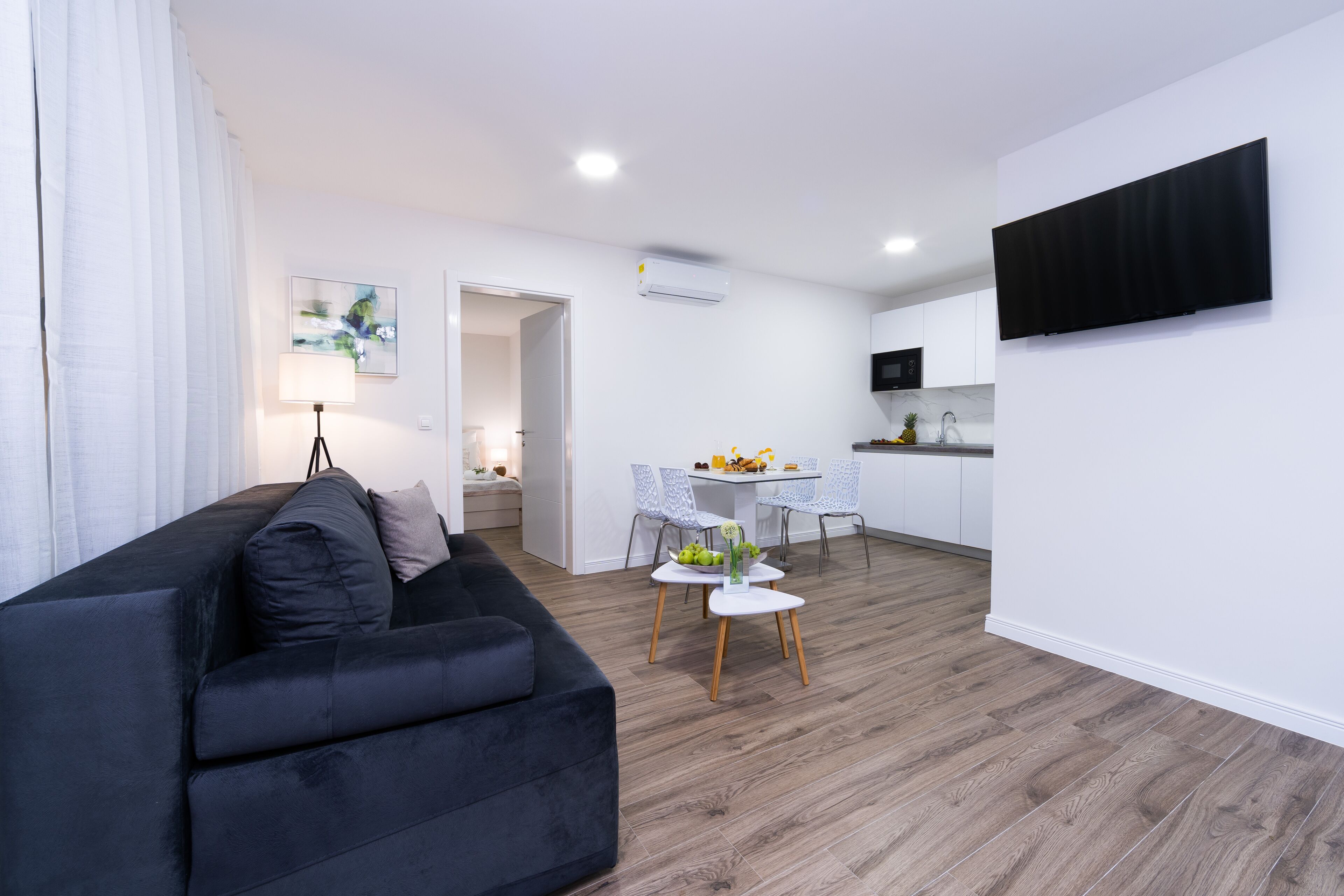 A modern living room with a black sofa, a dining area with a small table and chairs, and a kitchen with white cabinets and a flat-screen TV.