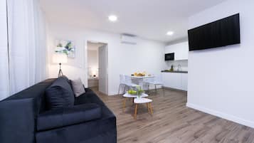 A modern living room with a black sofa, a dining area with a small table and chairs, and a kitchen with white cabinets and a flat-screen TV.