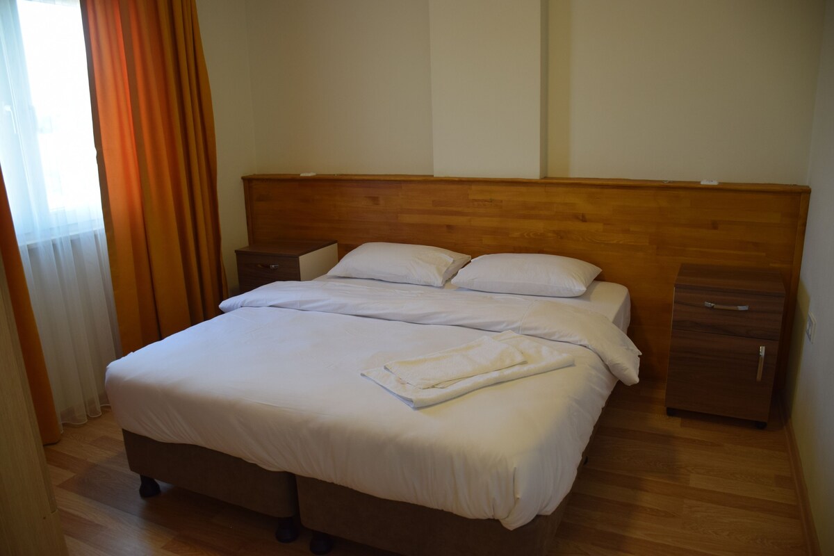Una cama bien hecha con sábanas blancas, cabecera de madera, una mesita de noche y un mueble con cajones de madera.