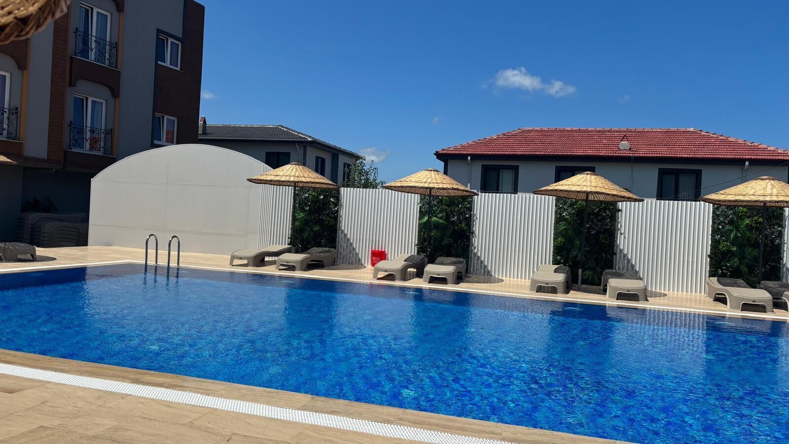 Una piscina con sillas reclinables y sombrillas, rodeada por una valla blanca y edificios al fondo.