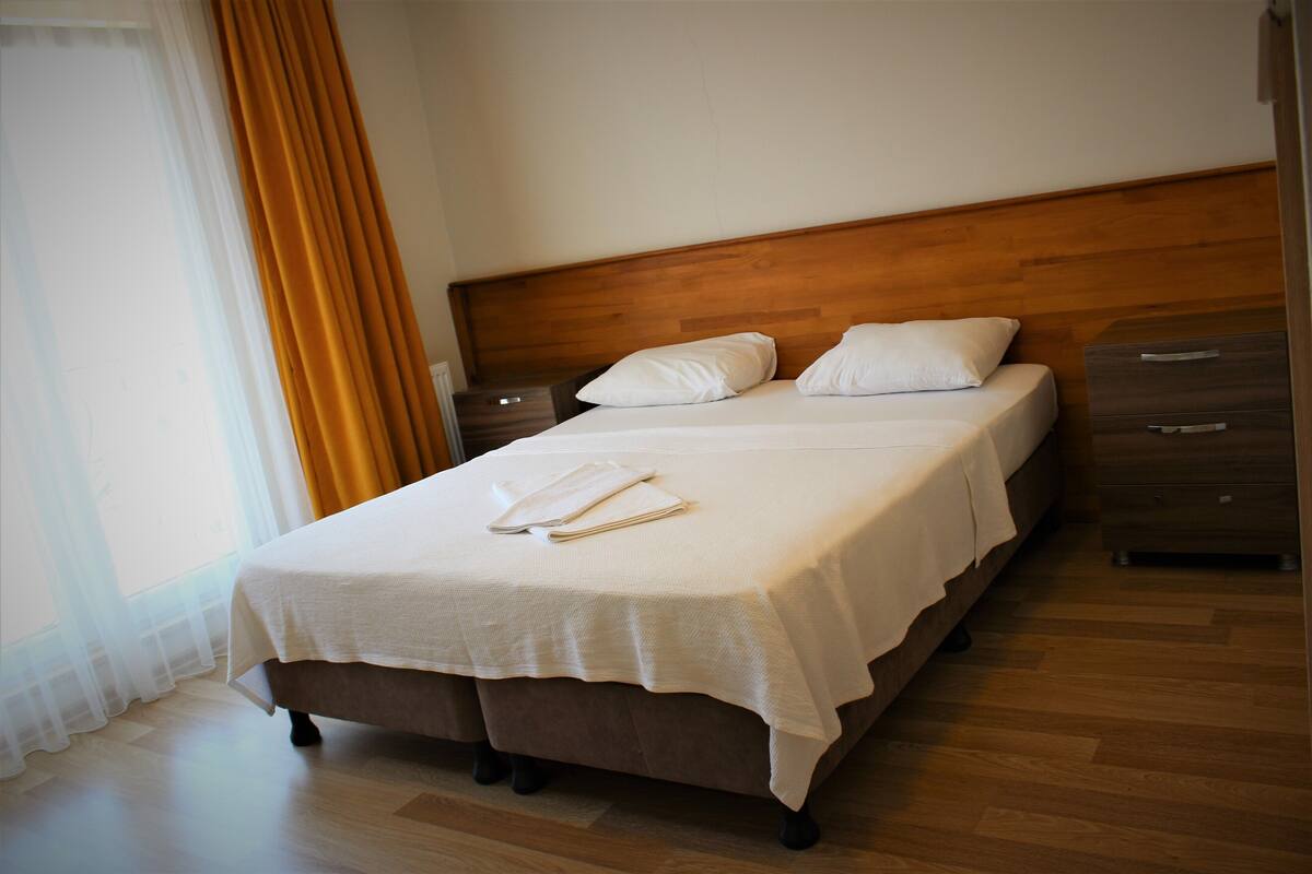 A neatly made bed with a wooden headboard, a bedside table with a lamp, and a window with curtains.