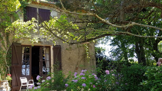 Un edificio rustico in pietra con una porta e delle finestre in legno, immerso in un rigoglioso verde e fiori colorati.