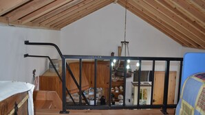 Une pièce aux hauts plafonds, avec des poutres en bois, une rambarde en métal noir et une vue sur un espace cuisine doté de meubles en bois et d’un luminaire suspendu.