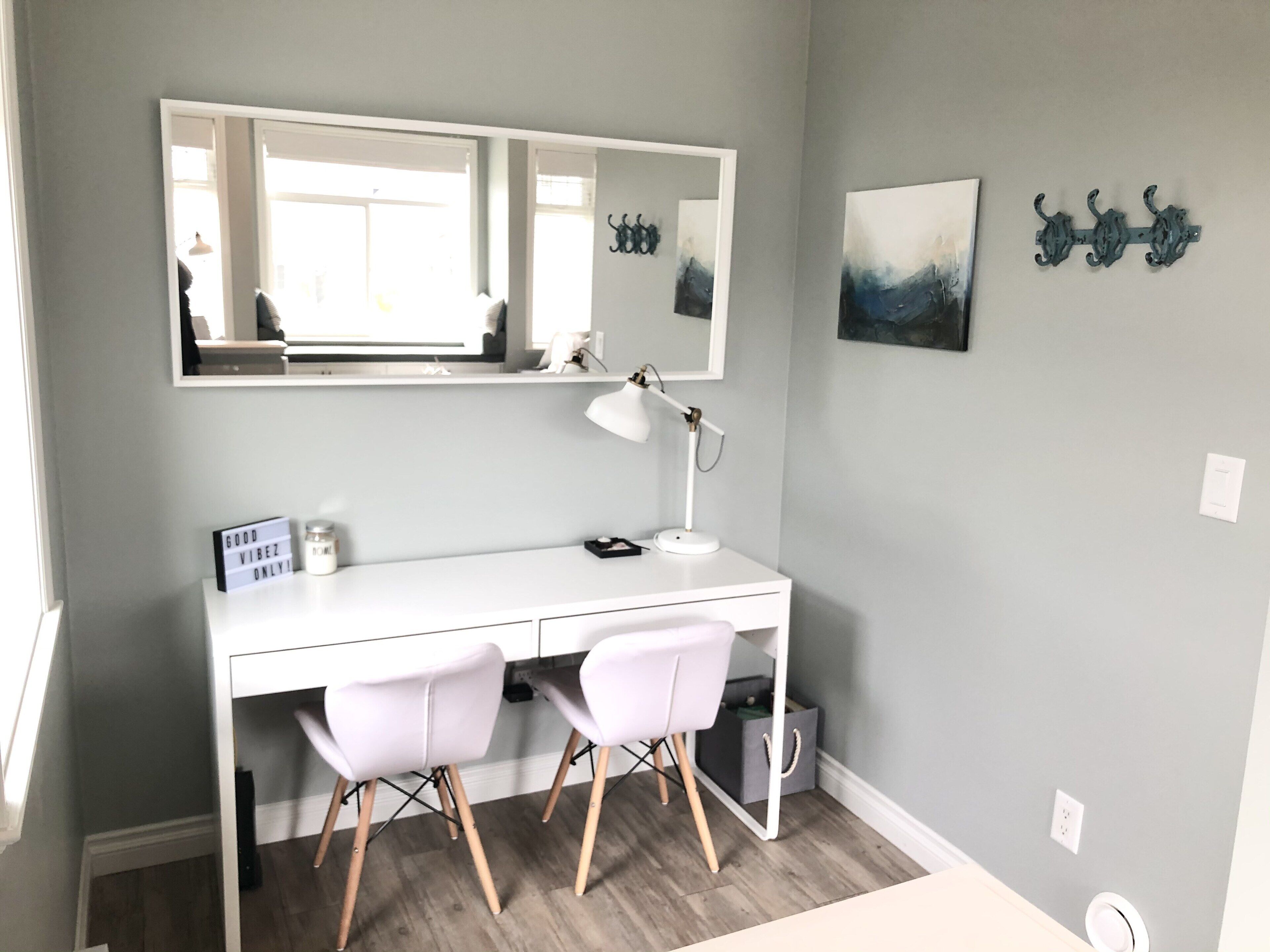 A white desk with two white chairs, a desk lamp, a mirror, and a small framed picture on the wall.