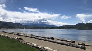 A lakeside view with mountains in the background, calm water, and a sandy beach with logs.