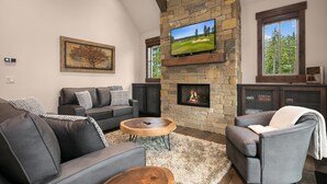 A living room with a stone fireplace, a television mounted above it, and a large window with a view of trees. - Big Home with LOW RATES! In the Heart of Suncadia Resort | Hot Tub | WiFi (Cle Elum)