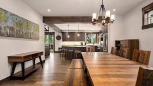 A spacious dining area with a long wooden table and benches, a kitchen with dark wooden cabinets, and a large painting on the wall. - Big Home with LOW RATES! In the Heart of Suncadia Resort | Hot Tub | WiFi (Cle Elum)