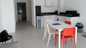 Een compacte keuken met witte kastjes, een magnetron en een kleine koelkast. Er staat een eettafel met twee stoelen, één rood en één wit. Een zwarte tas ligt op een klein tafeltje naast de stoelen.