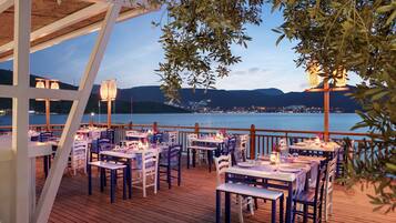 An outdoor dining area with tables and chairs, overlooking a body of water and a distant cityscape.
