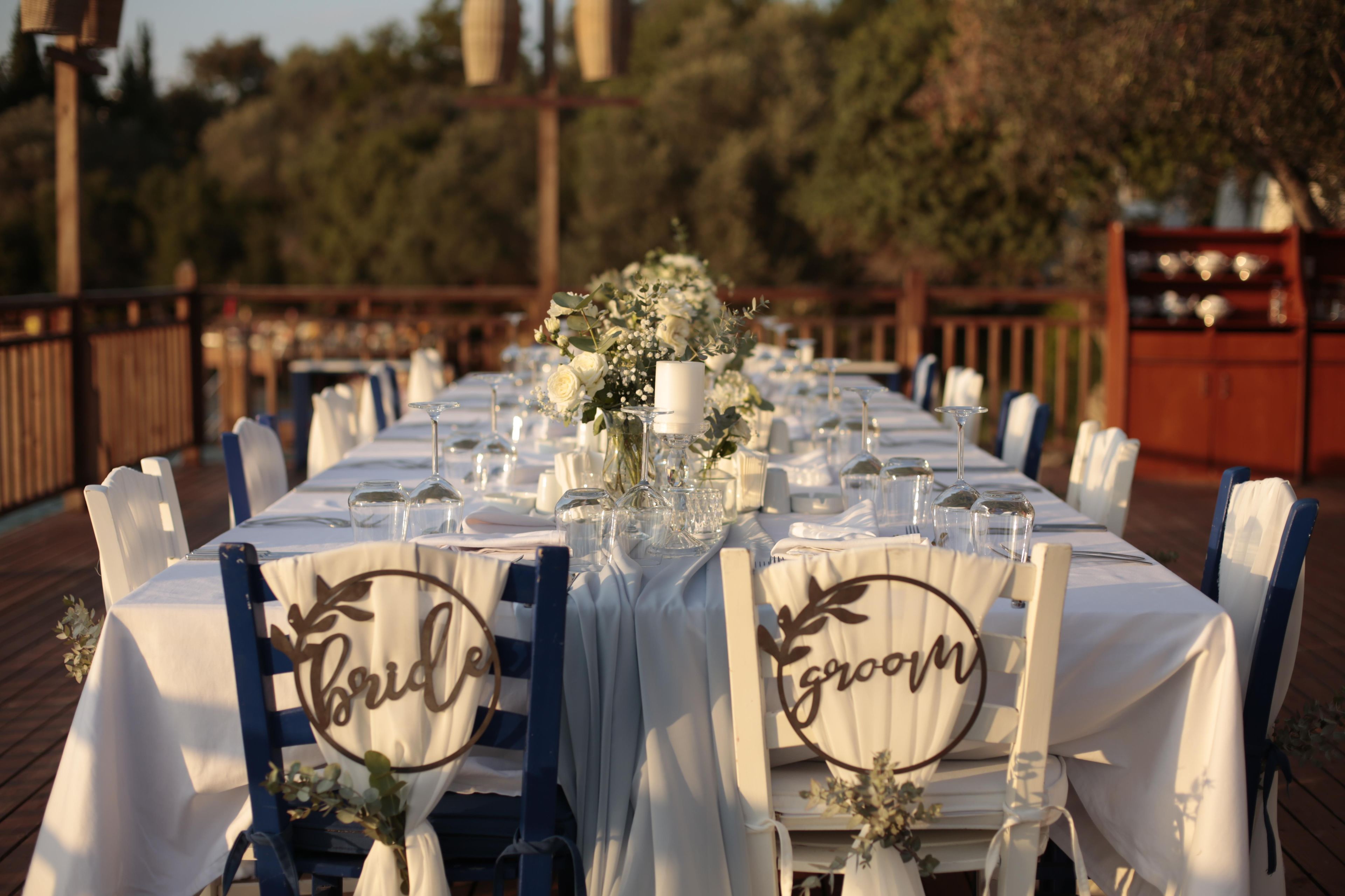 Una larga mesa de comedor dispuesta para una recepción de boda, con manteles blancos, cristalería y centros de mesa florales.